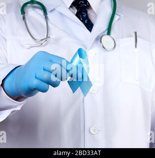 medico in un cappotto bianco e cravatta si erge e tiene un nastro di seta blu in un anello, indossando guanti medici sterili blu, simbolo della lotta e del trattamento o Foto Stock