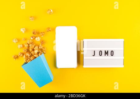 Abbreviazione parola JOMO scritto su un bordo decorativo su uno sfondo giallo brillante. Accanto alla schermata di simulazione dello smartphone. Vista dall'alto, disposizione piatta. Foto Stock