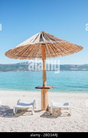 Ombrellone estivo in paglia e due lettini sulla spiaggia vuota della Croazia. Foto Stock