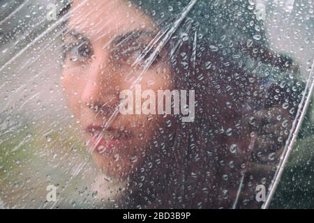 Triste volto di una ragazza dietro ombrello trasparente con pioggia. Primo piano Foto Stock