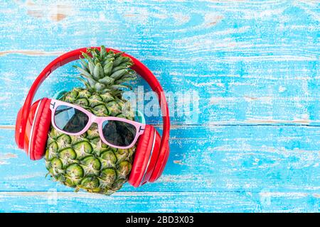 Estate e sfondo di viaggio da ananas indossando occhiali da sole e cuffie su bordo di legno blu con spazio libero. Foto Stock