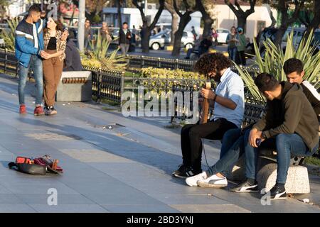 Giovane uomo con capelli ricci sulla strada sta cantando con uno strumento di violino. Si siede sul banco. La gente dà i soldi. Foto Stock