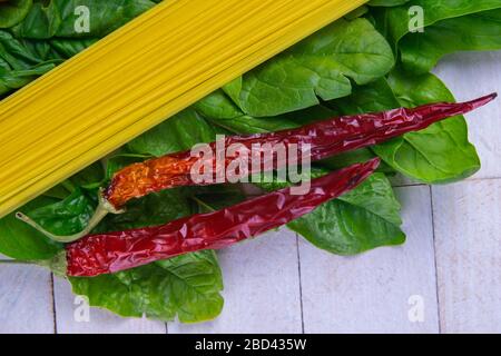 Spaghetti crudi e peperoni rossi caldi si trovano su un tavolo di legno bianco su foglie di spinaci Foto Stock