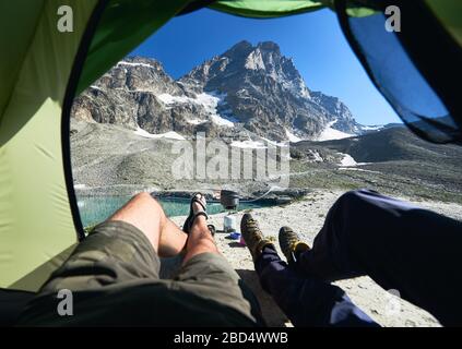 Gambe maschili all'interno della tenda da campeggio con montagna innevata sullo sfondo. Due viaggiatori che si trovano all'interno della tenda turistica e godono della vista delle splendide colline rocciose delle Alpi. Concetto di viaggio, trekking e campeggio. Foto Stock