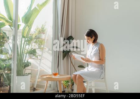 Giovane affascinante donna seduta su sedia e libro di lettura accanto alla finestra a casa. Foto Stock