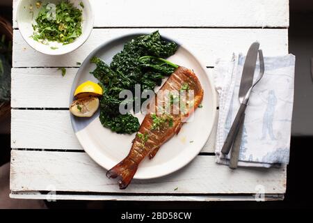 Un piatto con trota, kale e limone cotti su un tavolo di legno bianco con posate e tovagliolo Foto Stock