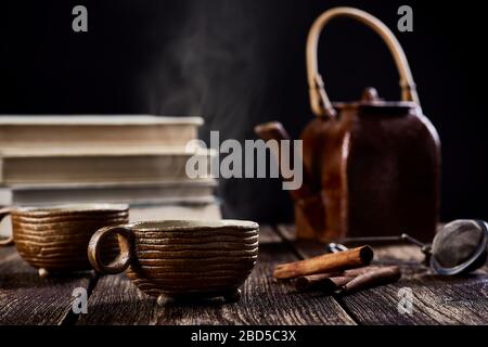 vista da primo piano della vita con tazza di tè o caffè in ceramica, teiera e cannella. Vecchio tavolo in legno, fumo di fronte ai libri e bac nero sfocato Foto Stock