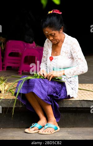 Ritratto verticale di una signora che tessere decorazioni tradizionali da foglie di palma in un tempio a Bali, Indonesia. Foto Stock