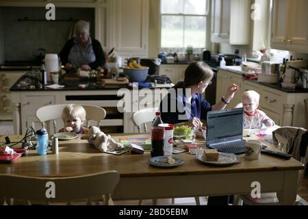 Madre che alimenta i suoi figli cena al tavolo da cucina mentre lavora da casa durante il periodo di autoisolamento - 2020 COVID-19 coronavirus pandemic Foto Stock