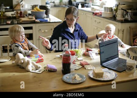 Madre che alimenta i suoi figli cena al tavolo da cucina mentre lavora da casa durante il periodo di autoisolamento - 2020 COVID-19 coronavirus pandemic Foto Stock