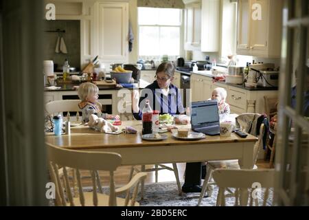 Madre che alimenta i suoi figli cena al tavolo da cucina mentre lavora da casa durante il periodo di autoisolamento - 2020 COVID-19 coronavirus pandemic Foto Stock