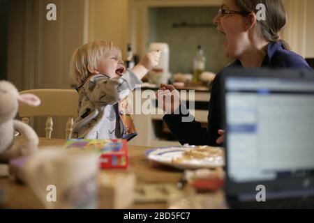 Madre che alimenta i suoi figli cena al tavolo da cucina mentre lavora da casa durante il periodo di autoisolamento - 2020 COVID-19 coronavirus pandemic Foto Stock