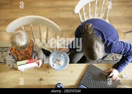 Madre che alimenta i suoi figli cena al tavolo da cucina mentre lavora da casa durante il periodo di autoisolamento - 2020 COVID-19 coronavirus pandemic Foto Stock