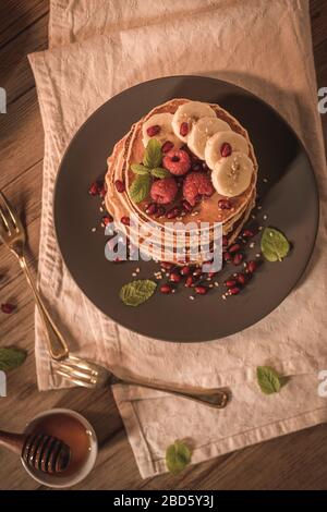 Pancake con lamponi, fette di banana, semi di melograno e miele su tavola d'annata in legno. Foto Stock