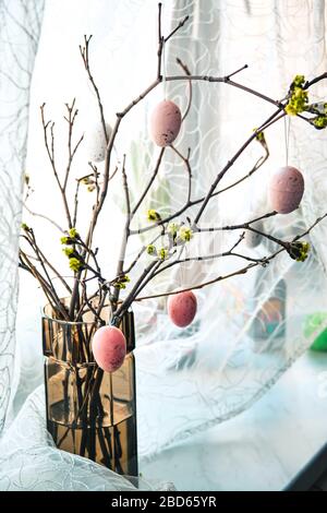 Interni domestici con decorazioni pasquali, rami freschi con boccioli verdi in vaso e uova color rosa pastello su uno sfondo chiaro di una finestra con tende. Foto Stock