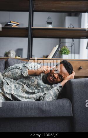 Stressato soldato afro-americano in uniforme militare piangendo mentre si parla su smartphone e soffrendo di PTSD a casa Foto Stock