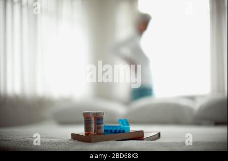 Due flaconi per pillole, un dispenser di farmaci e due libri seduti su un letto con una donna in piedi di fronte a una finestra sullo sfondo. Foto Stock