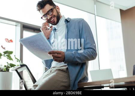 Vista ad angolo basso del lavoratore IT che tiene carta, parla sullo smartphone e sorride in uno spazio di lavoro Foto Stock