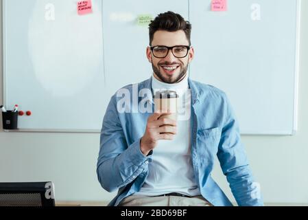 Maestro di Mischia che guarda la macchina fotografica e sorridente con una tazza di carta di caffè vicino al tabellone bianco con adesivi Foto Stock