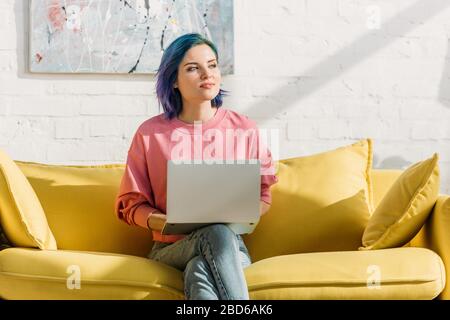 Freelancer con capelli colorati e notebook che guarda lontano e seduto su divano giallo con gambe incrociate in soggiorno Foto Stock