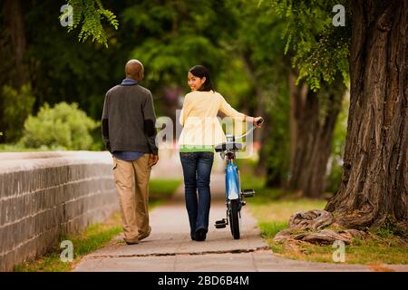 Giovane donna guarda sopra la spalla e sorride e spinge una bicicletta lungo come lei cammina lungo una strada residenziale con un giovane uomo. Foto Stock