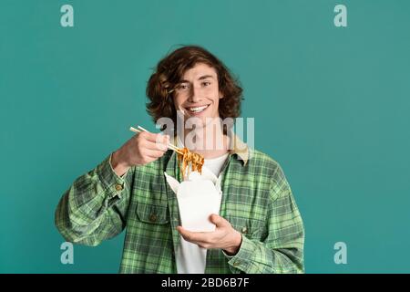 Cibo da asporto. Ragazzo felice millenario mangiare noodles cinesi da scatola usa e getta, sfondo turchese. Spazio vuoto Foto Stock