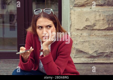 Ragazza triste in attesa di una chiamata o di un messaggio cellulare da parte del suo ragazzo seduto in una panchina all'aperto in strada con uno sfondo urbano Foto Stock