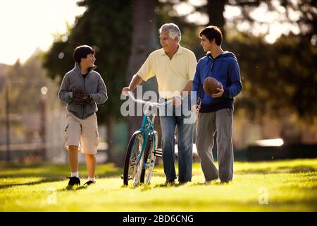 Sorridente uomo senior divertirsi a piedi con i suoi due nipoti attraverso un parco mentre wheeling una bicicletta. Foto Stock