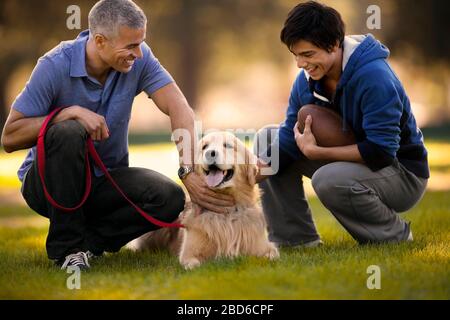Uomo maturo e suo figlio che si divertono a cucire insieme il loro cane mentre si accovacciano in un parco. Foto Stock