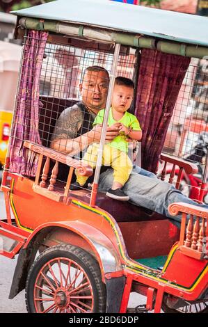 Un padre tiene il suo bambino mentre siedono nella parte posteriore di un risciò tradizionale e fanno il loro senso attraverso le strade trafficate di Phnom Penh. Foto Stock