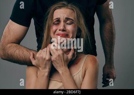 Il marito arrabbiato nella t-shirt nera è la ragazza bionda spaventata strangling nella camicia del undershirt beige con i capelli cuusciti ed i lividi sulla sua faccia. Posa su studio grigio b Foto Stock