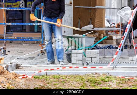 Posando le lastre di pavimentazione, un lavoratore con una pala da costruzione raccoglie una miscela di sabbia e cemento per livellare la fondazione. Foto Stock