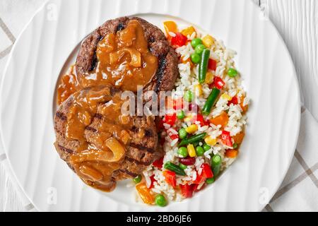 Bistecche di hamburger grigliate con sugo di cipolla e riso mescolate con verdure su un piatto bianco su un tavolo di legno, piatto, primo piano Foto Stock