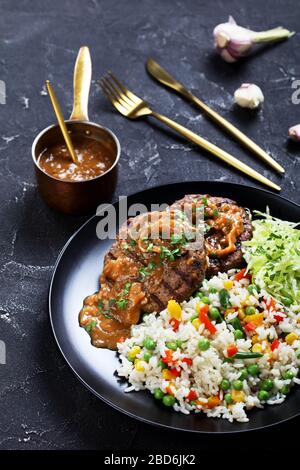 primo piano di una porzione di bistecche di manzo macinate alla griglia con sugo di cipolla, coleslaw e riso mescolati con verdure su un piatto nero su un tavolo di cemento, ve Foto Stock