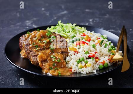 primo piano di una porzione di bistecche di manzo macinate alla griglia con sugo di cipolla, coleslaw e riso mescolati con verdure su un piatto nero su un tavolo di cemento, ho Foto Stock