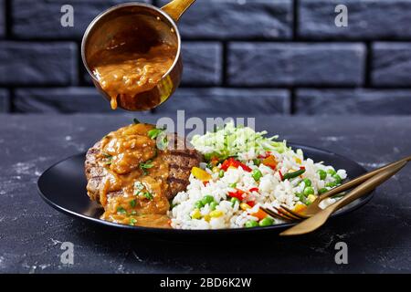 primo piano di una porzione di bistecche di manzo macinate alla griglia è stato versato con sugo di cipolla, coleslaw e riso mescolato con verdure su un piatto nero su una c. Foto Stock