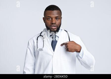 Confuso medico maschile nero in uniforme bianca che punta a se stesso Foto Stock