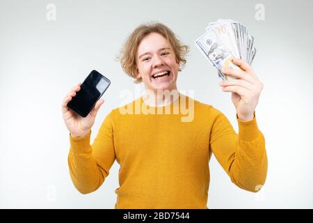 il ragazzo biondo felice tiene il suo stipendio e smartphone con un mockup su uno sfondo bianco Foto Stock