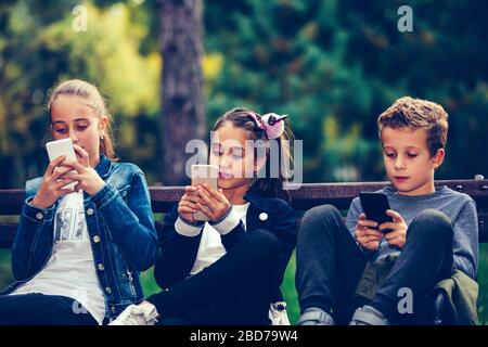 Gruppo di amici che giocano a videogame su smartphone dopo la scuola mentre si siede sul banco del parco. Foto Stock