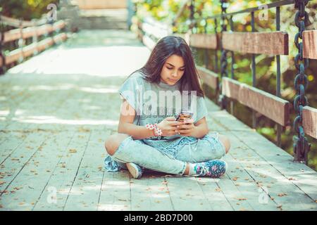 Donna infelice che testurla. Closeup ritratto sconvolto scettico donna seria chattando messaggi sul telefono dispiaciuti con conversazione seduta su un ponte legnoso Foto Stock