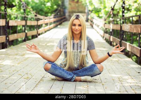 Giovane modello grazioso in abito casual seduta con gambe incrociate e mani in meditazione posa sul ponte nel parco estivo guardando la fotocamera. Foto Stock