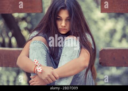 Giovane ragazza casual con lunghi capelli marroni seduti da soli sul ponte e abbracciando le ginocchia cercando solitario e premuroso. Foto Stock