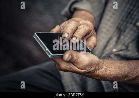 Anziani e tecnologia. Uomo anziano che utilizza il telefono isolato sfondo nero scuro. Closeup immagine ritagliata mani stropicciate che tengono il cellulare. Passato e futuro Foto Stock