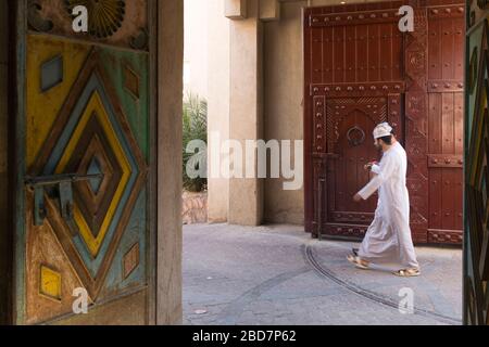 Uomini Omani in dishasdama camminare accanto ornati e decorativi tradizionali porte nel suk antico di Nizwa in Oman Foto Stock