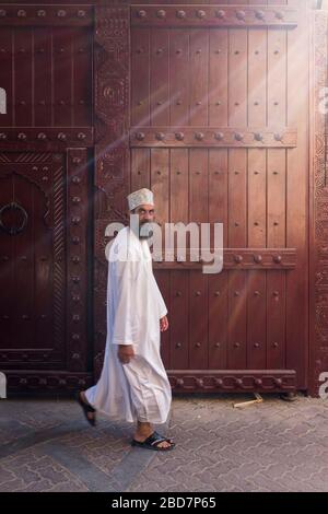 Uomini Omani in dishasdama camminare accanto ornati e decorativi tradizionali porte nel suk antico di Nizwa in Oman Foto Stock