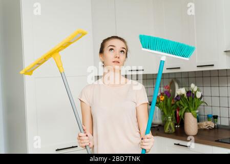 Una donna bella tiene un mop e una spazzola per la pulizia e il mopping nelle sue mani e sospiri di stanchezza. Una casalinga si trova in cucina e asciuga il sudore dal suo viso. Foto Stock