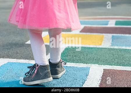 Primo piano delle gambe della ragazza in un abito rosa e hopscotch disegnato su asfalto. Bambini che giocano a hopscotch nel parco giochi all'aperto in una giornata di sole. All'aperto Foto Stock