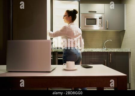 Una giovane donna che lavora con un notebook guarda nel frigorifero Foto Stock