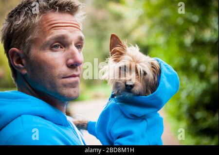 Il proprietario del cane che porta il suo piccolo amico migliore scruffy in felpa di felpa di felpa di felpa di felpa di felpa di felpa blu di corrispondenza che si levano all'aperto davanti al verde Foto Stock