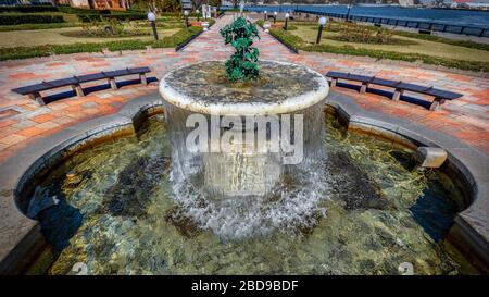 Il sole del mattino splende su una fontana in un parco con navi giapponesi e statunitensi sullo sfondo a Yokosuka, Giappone. Foto Stock